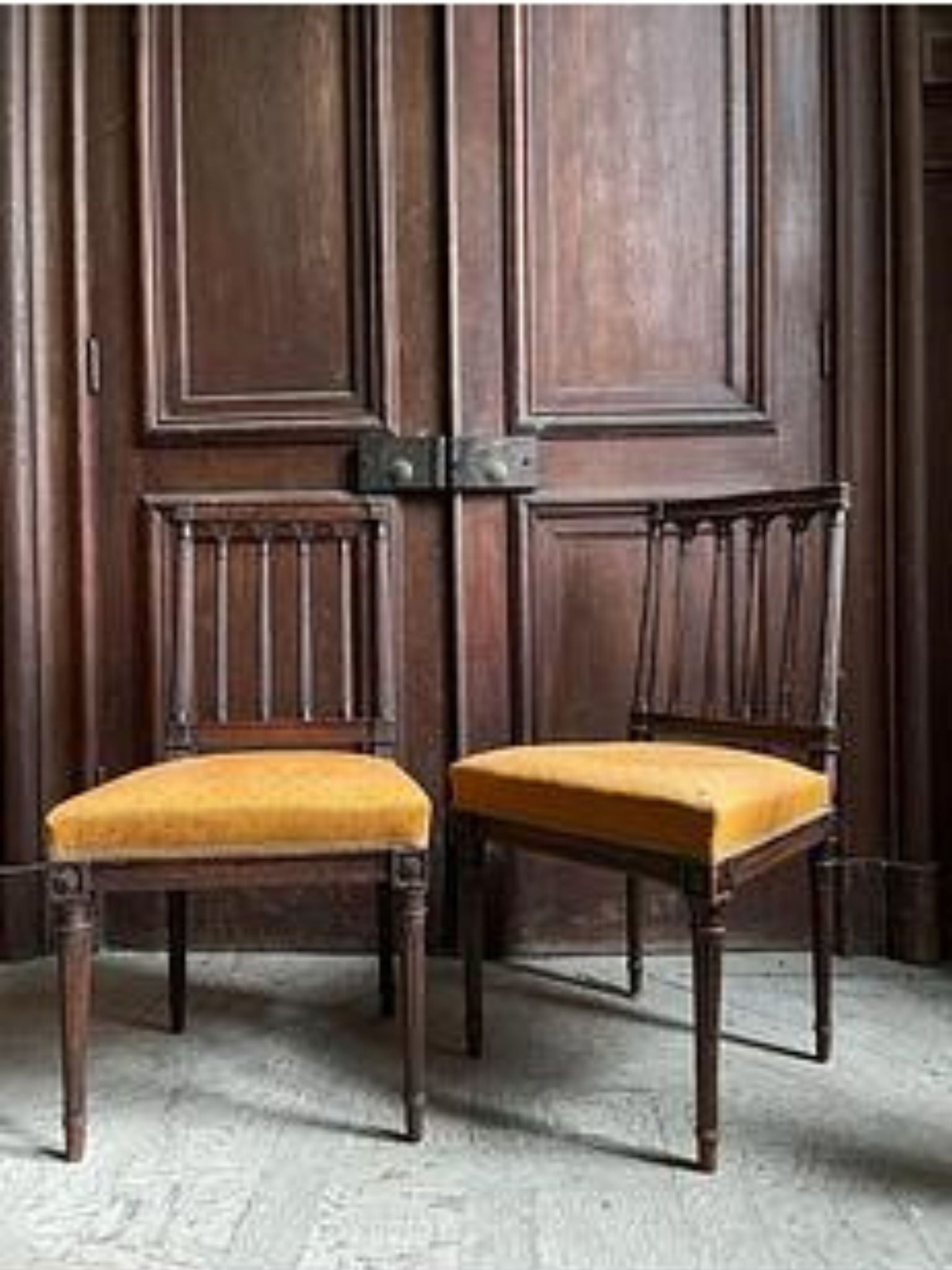 Two wooden chairs with yellow cushions in front of a large wooden door.